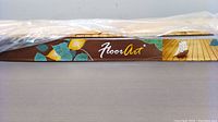 Photo of FloorArt packaging strip with brand logo and partial product image showing wood flooring installation on a deck.