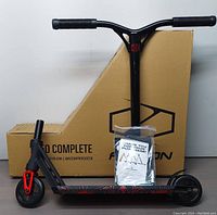 Assembled scooter front and side view against original packaging, including hardware bag.
