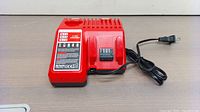 Red Milwaukee M18 & M12 multi-voltage battery charger with power cord, displayed flat on a surface, showing front and side with charging indicators and vents.