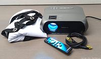 Photo showing Monster S5 projector with lens projecting blue light, folded white fabric screen with black edging, remote control, and HDMI cable.