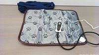 Full view of gray pet heating pad with black paw prints, brown trim, and digital temperature controller with attached cords