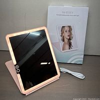 Front view of illuminated vanity mirror with pink frame, showing touch control buttons, USB cable, and product box in background.