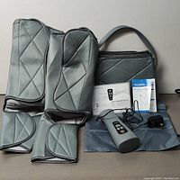 Full lot showing gray quilted leg wraps for thighs, calves, feet; handheld remote, power adapter, carrying case, and user manual on flat surface.