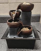 Front angle view of the small tabletop fountain showing water flowing between four brown cascading bowls supported by three grey stone pillars inside a black base reservoir.