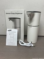Photo of the YIKHOM wireless sensor soap dispenser placed upright next to its retail box and manual, with a USB charging cable in front on a brown surface against a plain wall.