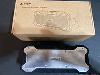 Front view of Aukey SoundTank Bluetooth speaker with silver grille and rubberized frame alongside its cardboard packaging box with line drawing of the speaker.
