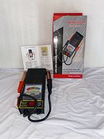Photo showing the battery tester laid in front of original packaging and a user manual.