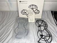 Two black plastic DJI Mini 3 Series 360 Degree propeller guard frames displayed on fabric surface, including original white packaging box, instruction manual, and foam protective insert.