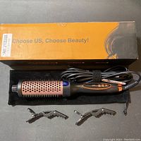 Hair straightener brush with attached power cord, two metal hair clips, and original yellow box packaging shown on a dark surface. The brush has a rose gold barrel with vents and a black handle with control buttons.