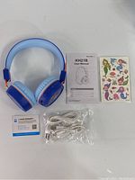 Kids blue and light blue headphones laid out with user manual, colorful stickers, warranty card, and extra cables in plastic bag.