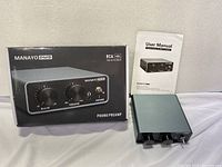 Manayo PM5 phono preamp device with original box and user manual displayed on white cloth surface. Front panel shows gain and volume knobs, power switch, and headphone jack.