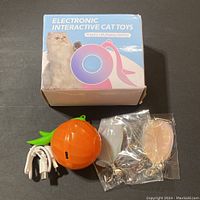 Image of the closed retail box with branding and the orange interactive cat toy with green leaf base, USB charging cable, and two sealed feather attachments.
