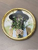 Front view of the round picture frame showing the gold textured border and a photo inside featuring a person holding wildflowers while wearing a black hat.