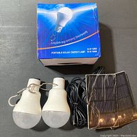 Two white LED bulbs with hooks, black solar panel with cable, and original box displayed on gray surface.