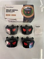 Four QueenMew Solar Animal Repeller units arranged in two rows along with the product box and instruction manual, showing front with red lights resembling eyes.
