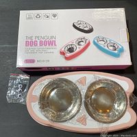 Photo of the Penguin Dog Bowl box with pink penguin-shaped feeding bowls inside, showing two stainless steel bowls inserted in the silicone base and a small bag with accessories.