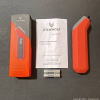 Ettenwolf T300 Plus Digital Tire Pressure Gauge in orange/gray with its packaging box, user manual, and three AAA batteries on a dark surface