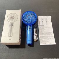 Photo showing blue mini handheld fan, its box with B8 model displayed, white USB charging cable, and user manual on a black surface.