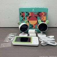 Full kit with original box, two baby cameras with green ear accents, video monitor, charging adapter, USB cable, mounting hardware.