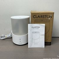 Front view of Clarston ultrasonic humidifier with power cord, manual, and original box next to it on a table.