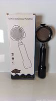 Photo showing the coffee bottomless portafilter with its box, the portafilter has a shiny metal frame and a black handle.