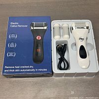 Photo of electric callus remover device in white with charging cable and two replacement roller heads in white plastic tray next to product box with product image and features.