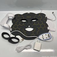 Black Hottoerak face mask with gold geometric pattern and eye/nose/mouth cutouts, white adjustable straps, lying flat with controller and power cables included