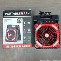 Photo of the portable two-in-one fan/light device beside its box showing the product and packaging with features: USB rechargeable, adjustable fan, LED light, built-in stand.