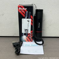 Black rechargeable Blukar LED torch with wrist strap, original box, user guide, and USB charging cable shown on grey surface.