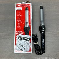 Revlon ceramic curling iron with power cord, original box, and instruction manual laid out on a flat surface. The curling iron has a silver barrel with black tips and clamp, black handle with Revlon logo.