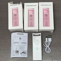 Set of three heated eyelash curlers, two in pink printed boxes and one out of box turned on. Includes two USB charging cables and instruction leaflets.