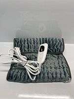 View showing folded heating pad with controller and power cord coiled and tied with a black twist tie on top, showing gray quilted soft material and wired remote control with black and white buttons.