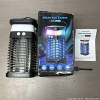 Photo showing the solar bug zapper unit together with its packaging box and instruction manual, placed on a wooden surface.