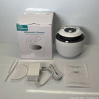 Photo showing the Ultrasonic Cleaner device, its retail box, power adapter, user manual, tweezers, and plastic holder.