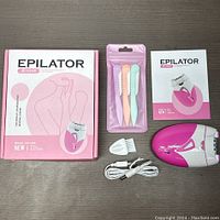 All items included in lot displayed on dark tabletop: Klend KD-189A epilator device, cleaning brush, USB charger cable, three pastel eyebrow razors, instruction manual, and box.