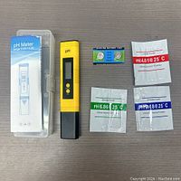 Top down view of digital pH meter in yellow with black lower electrode cover, calibration powders for pH 4.01, 6.86, and 9.18, 1.5V alkaline battery and clear plastic case with product label.