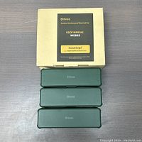 Three green outdoor weatherproof electrical boxes stacked on top of each other with the original packaging box and user manual in the background.