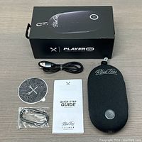 Top-down view of the speaker and all included accessories: Black fabric BlueTees PlayerGo magnetic speaker, USB charging cable, quick step guide booklet, black lanyard/strap in a plastic bag, and a circular adhesive sticker/decal. Black product box is seen in the background.