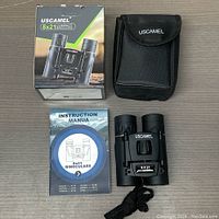 Photo of the boxed USCAMEL 8x21 compact binoculars alongside the binoculars themselves, the carrying case, and an instruction manual laid out on a flat surface.