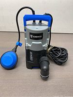 Front view of Deko pump showing black and grey body with blue handle and blue float switch, coiled power cord beside pump.