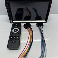 Stereo head unit with black remote control and two wiring harnesses with multi-colored wires and connectors laid out below it on a white surface.