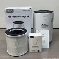 Kolkj air purifier main unit, HEPA filter, power adapter box, and instruction manual displayed on table with original box in background.