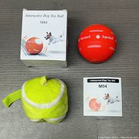 Red interactive dog toy ball with ‘Supreba II’ branding, alongside its white box featuring an image of the ball and a dog, a small instruction manual, and a plush tennis ball-style dog toy on a gray surface.