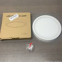 Top-down view showing LED ceiling lamp unit, original flat cardboard box with product illustration, and small bag of installation hardware.