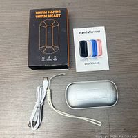 Top-down view of the hand warmer set showing the silver hand warmer device, white USB charging cable, user manual, and original box on a wooden surface.