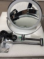 Front view of round LED vanity mirror with 10X magnification printed on mirror surface. Included USB charging cable and mounting hardware shown on white protective foam.