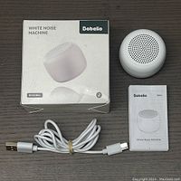 Photo showing the white noise machine with its original box, USB charging cable, and user manual placed next to each other on a wooden surface.