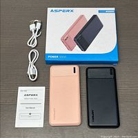 Two Asperx 10000mAh power banks side by side, peach and black colors, with two white USB cables, manual, and product box on a flat surface.