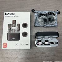 Lot contents showing packaging box, two wireless microphones in charging case, two grey windshields, USB charging cable, and grey storage pouch spread out on surface.