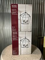 Photo of two boxed 4-light cage pendant lights by Home Decorators Collection in matte black finish. Boxes show the image and measurements of the lights, one box sealed, one opened but complete.